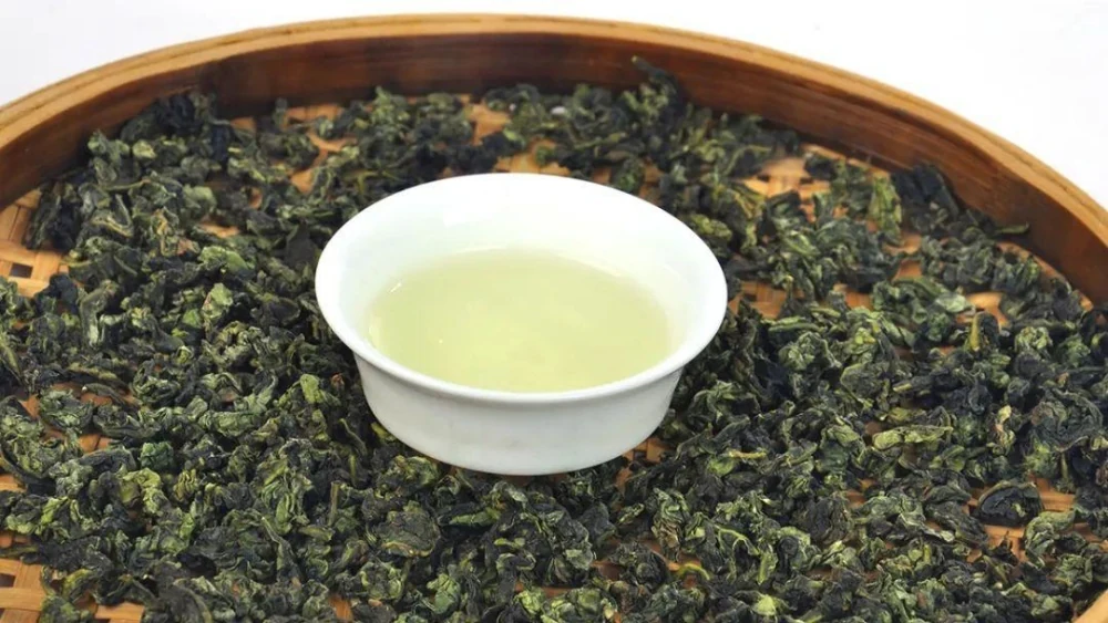 Una taza blanca de té claro descansa sobre una bandeja de bambú cubierta de hojas enrolladas de color verde oscuro.