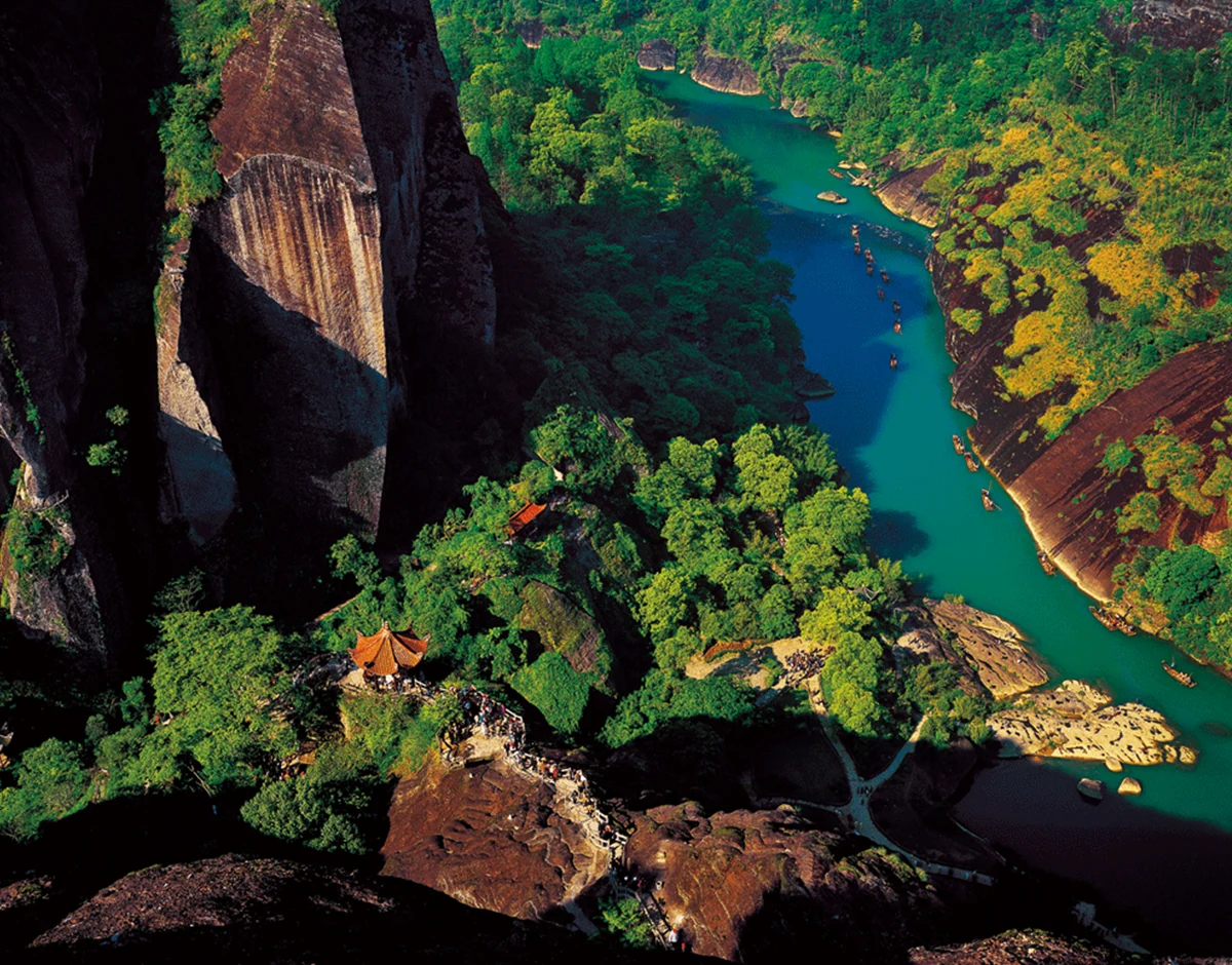 Une vue plongeante révèle une rivière bleu-vert avec de petits radeaux, des pavillons aux toits orange et des chemins sinueux.