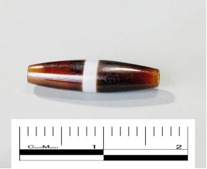 Une perle allongée brun-rouge à motif blanc en forme de croix, près d’une échelle centimétrique.