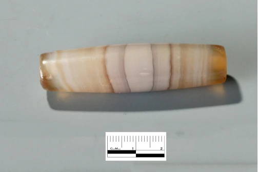 Une parure allongée en agate à bandes crème et brunes, près d’une échelle centimétrique.