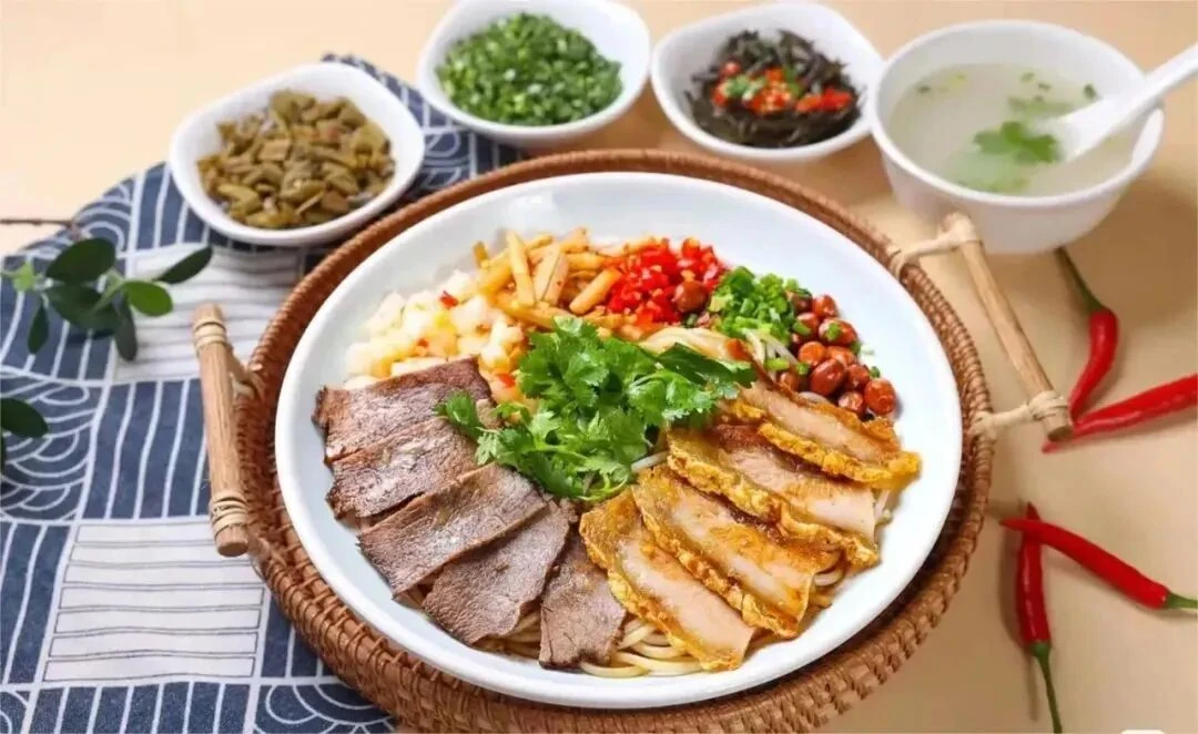 Миска рисовой лапши с ломтиками мяса, арахисом, рубленой зеленью и красным перцем чили; рядом приправы и суп.