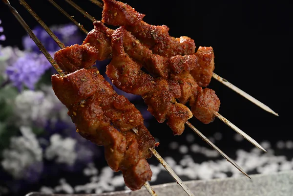 深色背景前，四根扁平的金屬籤上串著烤至褐色的肉，表面撒有芝麻與紅色調料。