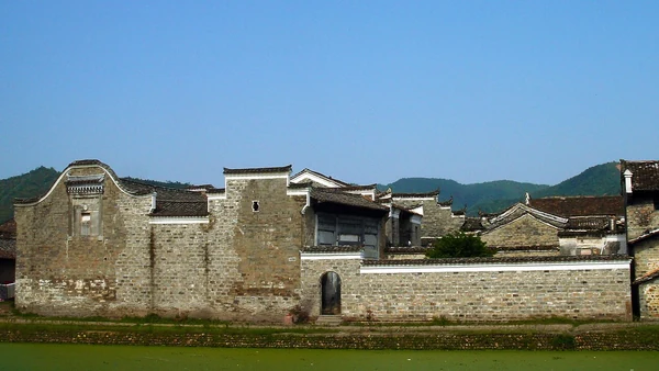 Une large maison de briques grises aux pignons étagés se dresse au bord de l’eau à Liukeng, devant des collines boisées.