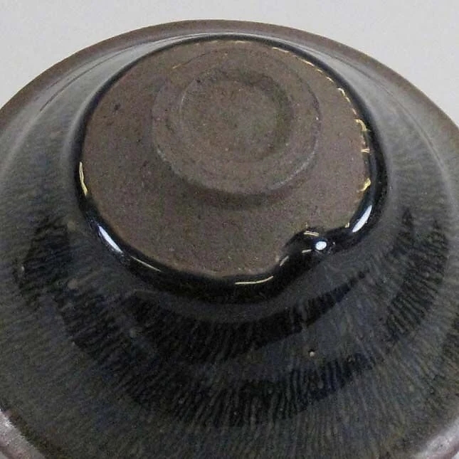 La parte inferior de un cuenco Jian, con un pie circular de arcilla descubierto y esmalte oscuro brillante.