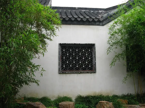 A decorative lattice window in a white garden wall between green bamboo plants at Geyuan.