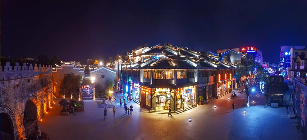 Illuminated shopfronts and pedestrians in Dongxi Xiang beside a stone wall at night.
