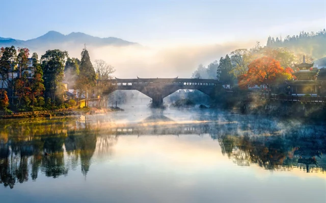 Eine überdachte Brücke führt zwischen Nebel und herbstlich gefärbten Bäumen über stilles Wasser.