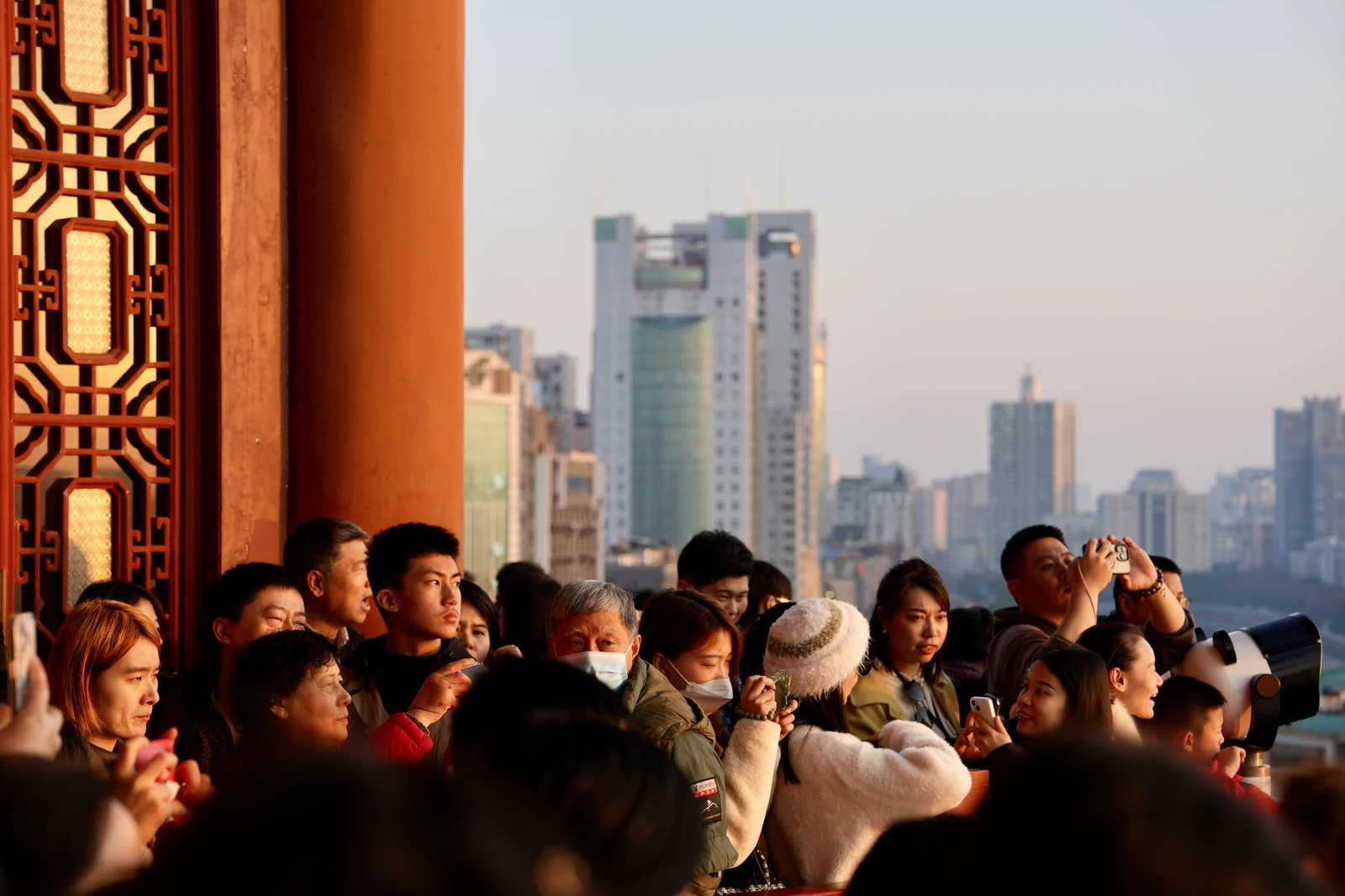 Des visiteurs près des colonnes rouges du pavillon Tengwang, avec les immeubles modernes de Nanchang au loin. Photographie de 2024.