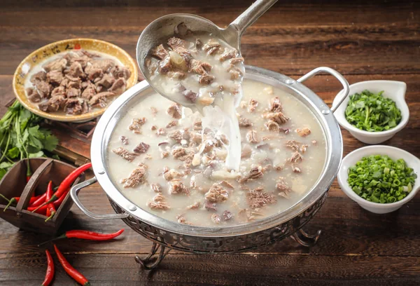 Un cucharón vierte caldo de aspecto lechoso y trozos de carne en una olla metálica poco profunda, con pequeños cuencos de hierbas picadas y chiles rojos sueltos cerca.