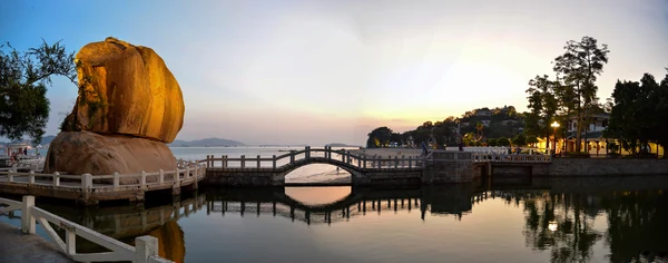 Uma ponte de pedra atravessa água tranquila junto a uma grande rocha, ao pôr do sol.