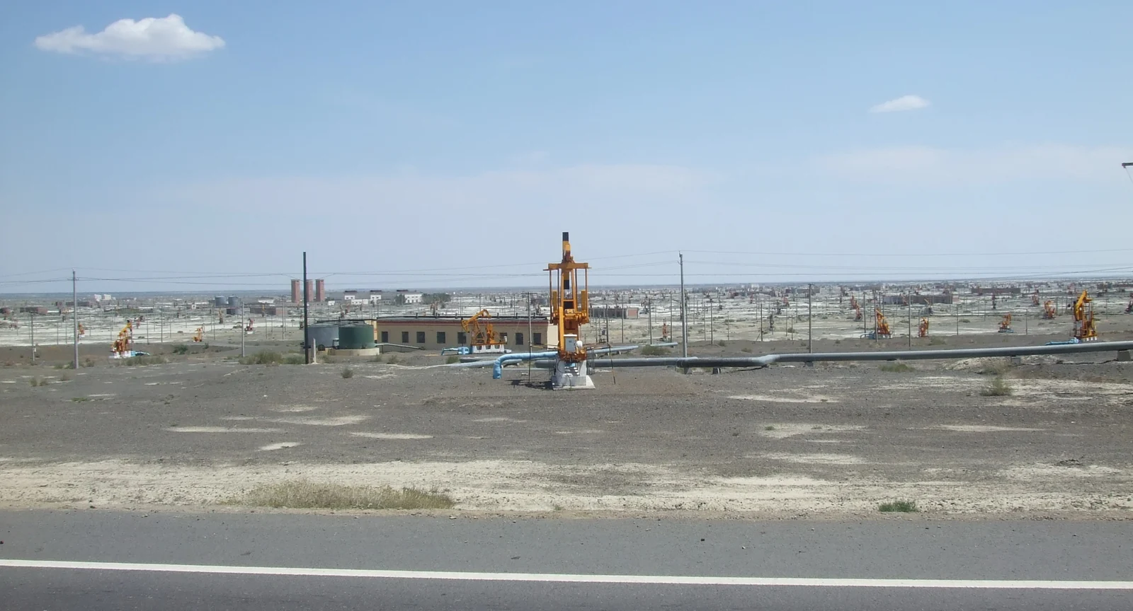 A pumpjack beside a road, with many more across the gravel plain and low buildings beyond.