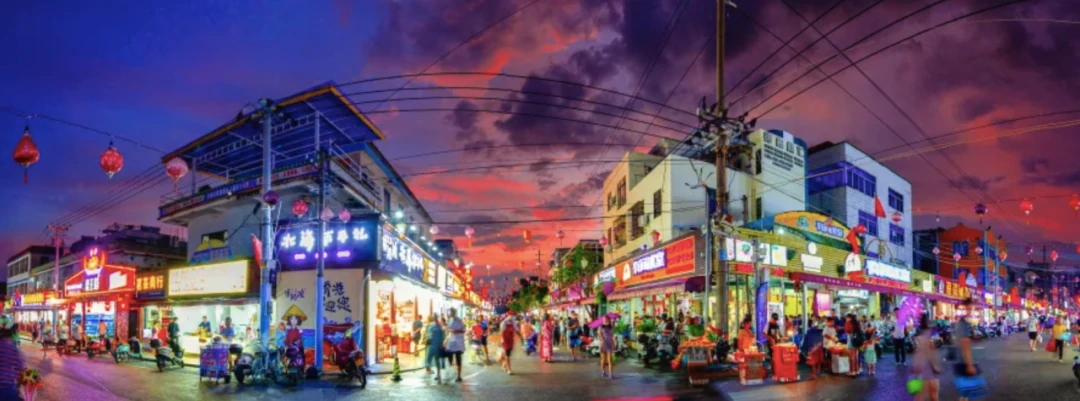 Personas y comercios de comida iluminados llenan una intersección de Qiaogang bajo un cielo vespertino de colores intensos.