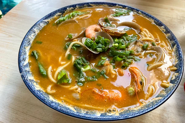 Un cuenco de caldo anaranjado con fideos, gambas, calamar y verduras.