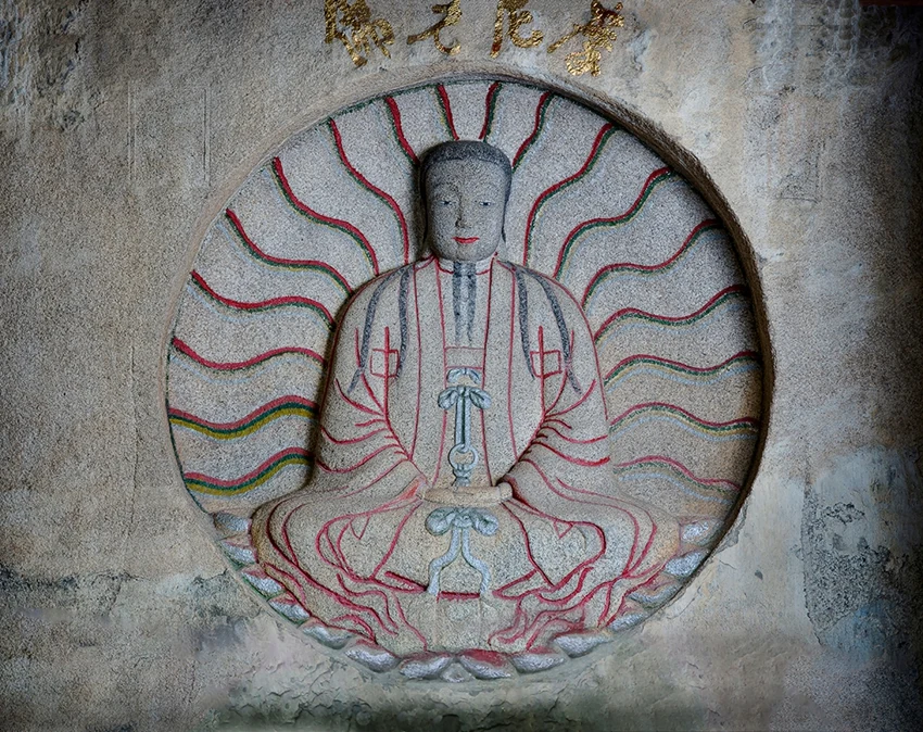 Une figure assise est sculptée dans une auréole circulaire dessinée par des lignes colorées rayonnantes.