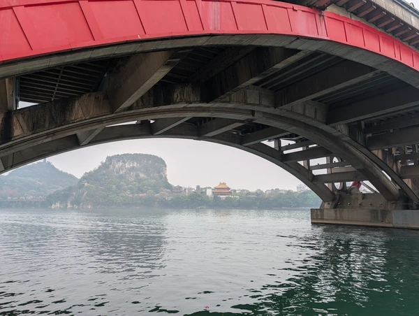 The underside of Wenhui Bridge frames the Liujiang riverbank and a wooded hill in Liuzhou.