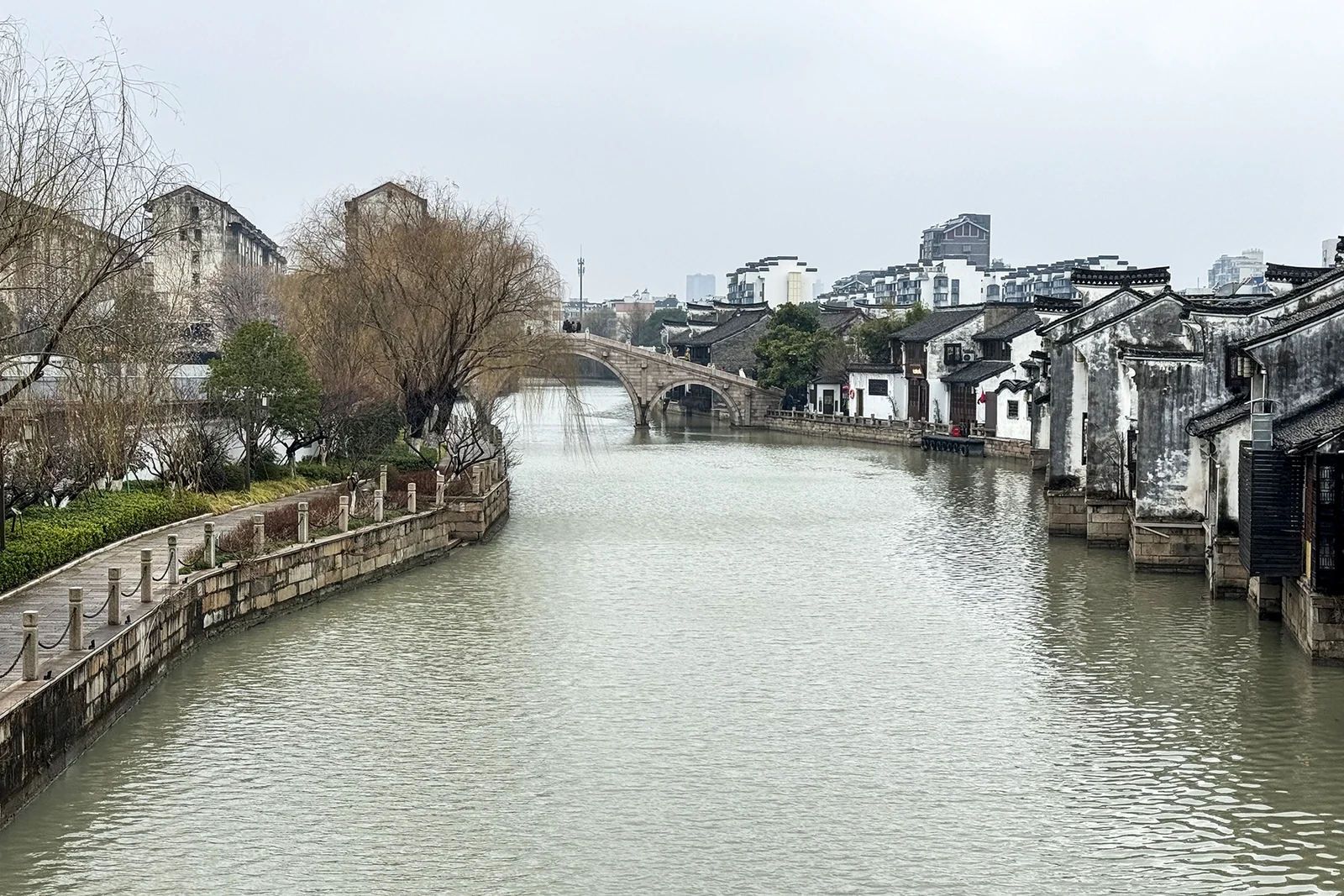 一座石橋橫跨嘉興大運河，旁邊是白牆建築和林蔭小路。