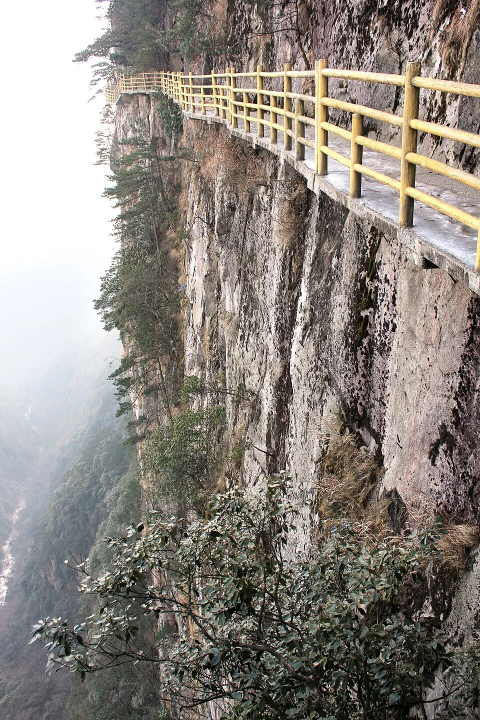 Une passerelle à rambarde jaune longe une falaise abrupte au-dessus d’une vallée boisée et brumeuse du mont Mingyue, à Yichun.