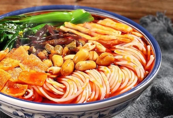 A bowl of rice noodles in red broth with peanuts, greens and golden fried toppings.
