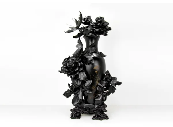 Eine glänzende schwarze geschnitzte Vase, umgeben von Blüten, Blättern und einem Schmetterling, steht auf einem verzierten Sockel vor weißem Hintergrund.