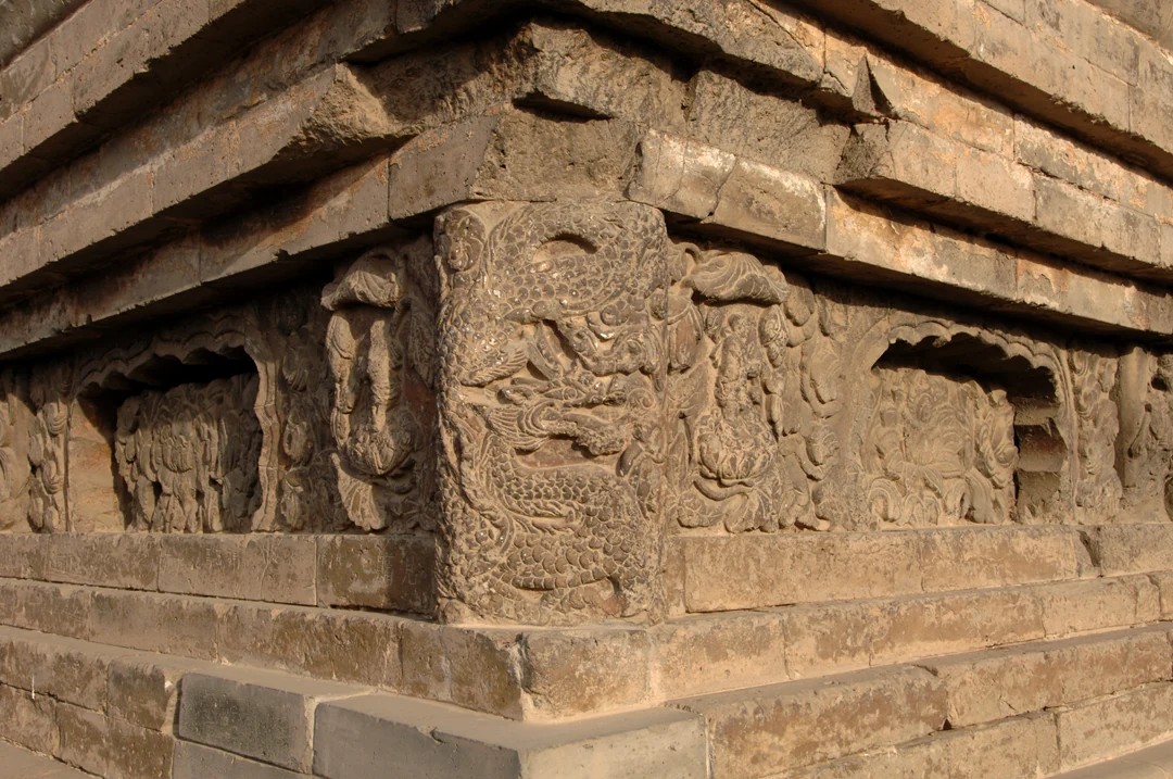 Figuras desgastadas pelo tempo e painéis ornamentais estão esculpidos em redor de um canto saliente da base de um pagode de tijolo.
