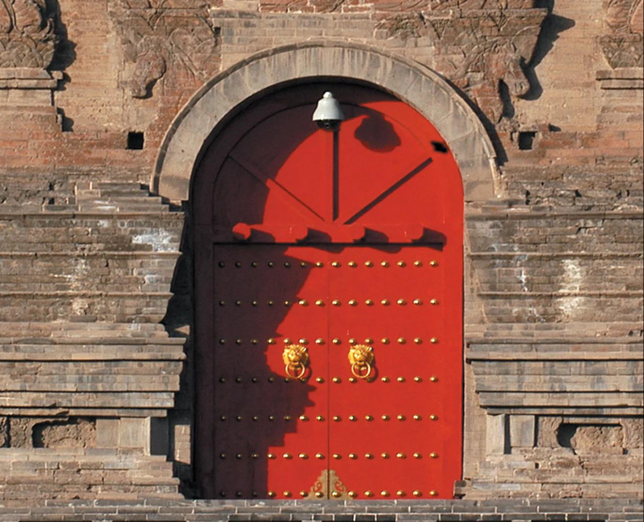 Uma porta dupla vermelha em arco, com tachas douradas e ferragens de argola, é enquadrada por alvenaria de tijolo e figuras esculpidas.