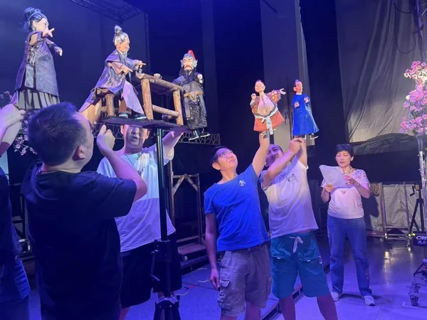 Performers raise small hand puppets above a stage while others watch.