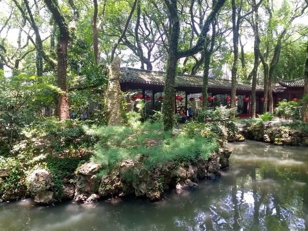 A tree-shaded covered timber walkway with red lanterns runs beside a pond in Tianyi Pavilion garden.