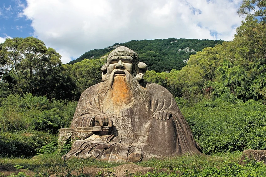Uma figura monumental sentada, esculpida em pedra, diante de uma encosta arborizada.