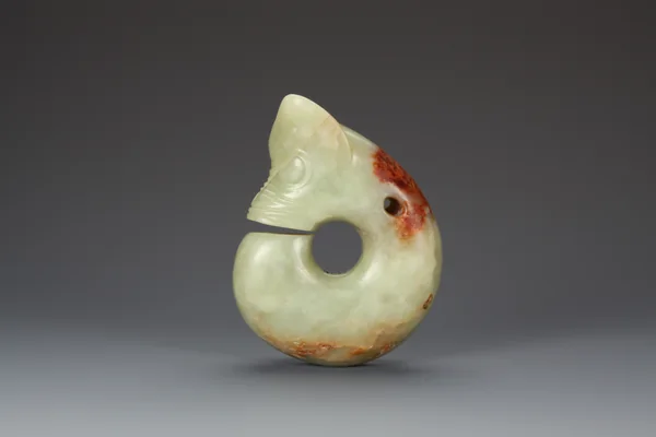 Une sculpture recourbée en jade vert pâle présente une grande ouverture circulaire, un étroit interstice au niveau du cou et des taches brunes à sa surface.